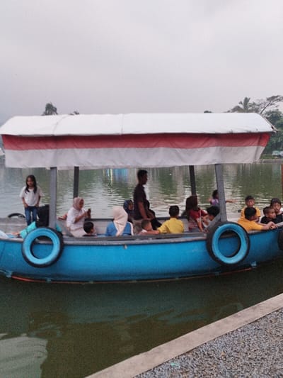 A blue passenger boat, featuring a white canopy with a red horizontal stripe, is positioned on a body of water adjacent to a concrete and gravel bank in Taman Sari, Indonesia. Approximately 15 individuals, comprising both adults and children, are on board. One young girl in a white shirt and blue jeans stands near the front-left of the vessel. Seated passengers include two women wearing hijabs, one of whom is observing a mobile phone, and several children, some in yellow shirts, are situated towards the right side of the boat. A male figure in a dark shirt is partially silhouetted in the center. Two black rubber tires are attached to the boat's exterior as fenders. The environment consists of calm, greenish water reflecting the overcast sky, with distant green trees visible along the far bank.