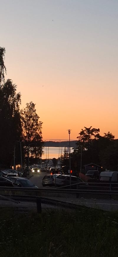 The image captures a sunset scene in Salpakangas, Finland. The sky transitions from a pale blue at the top to a warm orange glow near the horizon. In the foreground, a dark, grassy embankment leads down to a road bordered by a metal guardrail. Several cars are parked or driving on the road, with their headlights illuminated. Trees with dark foliage are visible on the left and right sides of the image, partially obscuring the view. In the background, a body of water, likely a lake or bay, reflects the sunset colors. Several sailboats are moored in the water, their masts silhouetted against the sky. A lamppost with a light fixture stands on the right side of the road. The overall impression is a peaceful evening setting, with a hint of urban life due to the presence of vehicles and infrastructure.