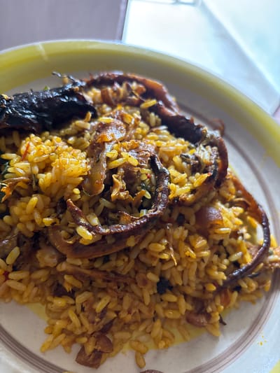A plate of jollof rice with fish is presented. The dish is plated on a yellow-rimmed plate. The jollof rice is a mix of golden-yellow rice, pieces of fish, and other ingredients. There are several pieces of fish that appear dark brown and somewhat crispy. The dish is situated in Rumuokwuta, Nigeria.