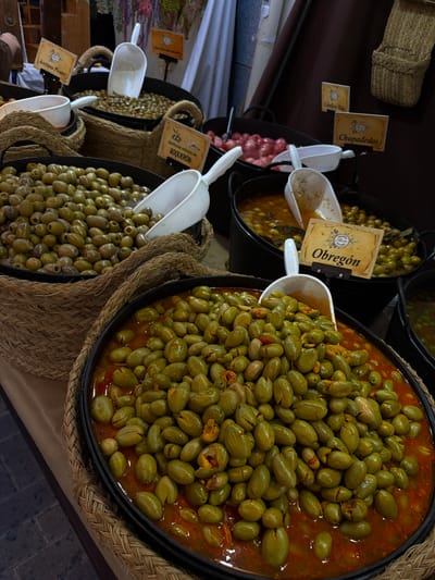 The image depicts a display of various types of olives in a market setting, presumably in Burjassot, Spain. Several black metal containers lined with woven baskets hold different varieties of olives, each with its own identifying sign. The olives vary in size, color, and preparation, some appearing whole, others pitted, and some submerged in liquid. White plastic scoops are placed in each container, presumably for customers to serve themselves. A basket in the upper left corner and a partially visible item in the back suggest a bustling market environment.