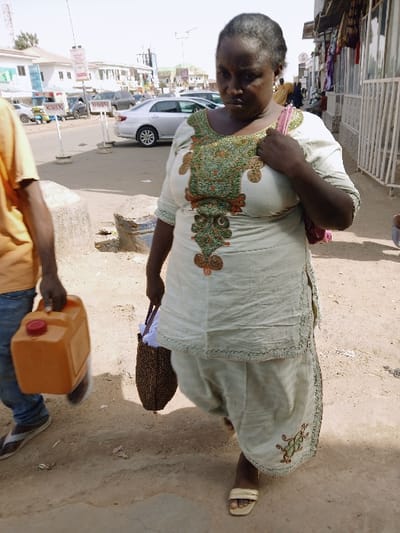 This image captures an outdoor street scene during bright daylight in an urban or semi-urban environment consistent with a town in Zambia. Kungulu School is not explicitly visible within the frame.
In the foreground, a dark-skinned, middle-aged woman walks towards the left side of the frame, her gaze directed downwards. She wears a light greenish-beige tunic top and matching loose-fitting trousers, both featuring intricate green, gold, and orange embroidery patterns. Her hair is dark with visible graying, pulled back. A pink or purple shoulder bag rests on her left shoulder, and she holds a brown, woven tote bag in her right hand. She wears light-colored open-toed sandals.
Adjacent and slightly to her left, a partially visible individual, shown from the mid-torso down, carries a rectangular orange plastic jerrycan with a red cap in their right hand. This individual wears an orange/tan t-shirt, blue jeans, and brown sandals or flip-flops, walking in the same direction.
The ground consists of an uneven, dry, light brown dirt path or sidewalk. In the mid-ground and background, a paved road with multiple vehicles, including a silver sedan and a blue/yellow three-wheeled vehicle, is visible. White, multi-story buildings line the street, some featuring storefronts with metal grilles and glass windows. Textiles are displayed outside a building on the right. Concrete bases for street poles are present on the sidewalk. The scene is illuminated by strong, direct sunlight.