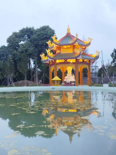 The image shows a pagoda reflected in a body of water, likely a pond or moat. The pagoda features a multi-tiered roof adorned with dragon ornamentation. It is illuminated from within, casting a warm yellow glow. Inside the pagoda, a white statue is visible. The surrounding environment includes trees and vegetation. The water is murky and contains floating debris. The photo may have been taken in the city of Phú An, Vietnam.