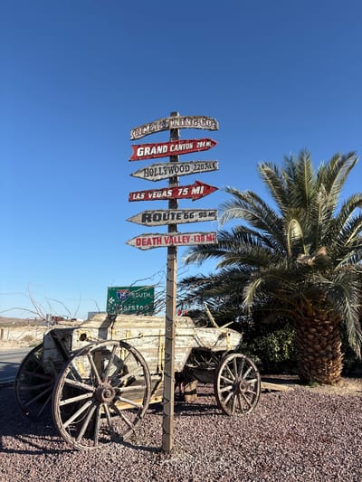 A directional signpost stands in an outdoor environment. The signpost is made of wood and has multiple signs pointing in different directions with place names and distances. From top to bottom, the signs read: "CIMARRON MINING CO.", "GRAND CANYON 284 MI", "HOLLYWOOD 320 MI", "LAS VEGAS 75 MI", "ROUTE 66 - 90 MI", and "DEATH VALLEY - 135 MI".
In the foreground, a weathered wooden wagon with large spoked wheels sits on a bed of gravel. Behind the wagon and to the right, a large palm tree with green fronds is visible. A green highway sign with white lettering indicating "I-15 SOUTH" and "Barstow" is partially visible behind the directional signpost. The background consists of a clear blue sky and a dry, arid landscape with sparse vegetation and a distant road. The overall scene appears to be in a desert environment. The location is Unknown, United States.