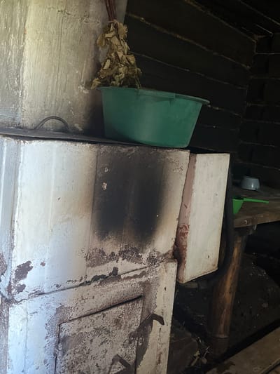 The image shows an interior view of what appears to be a rustic wooden structure, possibly a sauna or bathhouse, in Большой Гондыр, Russia. The focal point is a white, brick-like stove that is weathered and shows signs of soot staining. On top of the stove, a green plastic basin sits next to a bundle of dried leafy branches, likely a venik used in saunas. The background is dominated by dark, rough-hewn wooden walls. To the right of the stove, a wooden shelf holds two small basins, one blue and one green ladle. The overall impression is one of a functional, traditional space.
