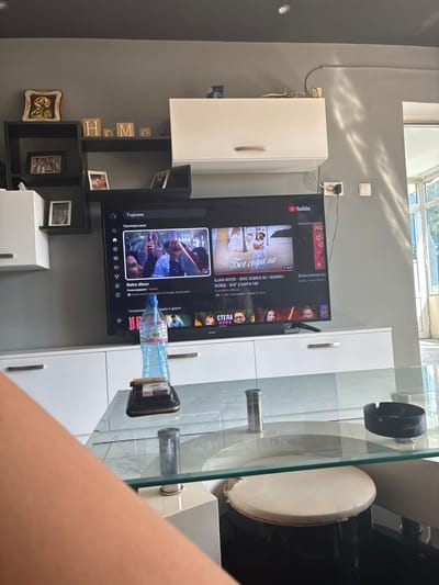The image depicts an indoor scene, likely a living room, with a prominent television screen displaying a YouTube video. The television is mounted on a white media console. On the wall above the console, decorative letters spelling "HOME" are visible, along with a framed religious icon and several framed photographs. Shelving units are attached to the wall, holding more framed pictures.

A glass-top coffee table is in the foreground, reflecting the objects placed on it and the surrounding environment. On the table, a water bottle, a wallet with cigarettes, and an ashtray are present. The overall ambiance suggests a casual, lived-in space.

The scene is consistent with residential interiors that might be found in the city of Radievo, Bulgaria.