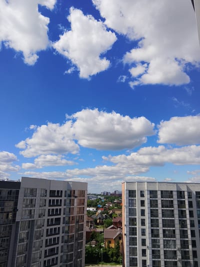 The image captures an aerial view of a residential area on a bright, cloudy day, likely in Zhilino-1, Russia. Two modern apartment buildings dominate the foreground, with their multi-pane windows reflecting the sky. The building on the left has a predominantly gray facade with darker vertical elements, while the one on the right features a lighter gray exterior with sections of windows tinted with a coppery hue.
Beyond the apartment buildings, a scattering of houses with pitched roofs, varying in color from brown and orange to white, are visible. These houses are interspersed with lush green trees, suggesting a mix of urban development and natural landscape. In the distance, a hazy cityscape is discernible, with more buildings of varying heights under the expansive blue sky filled with fluffy white clouds. The lighting suggests it is daytime, with sunlight illuminating the scene. There are no people or animals visible in the image.