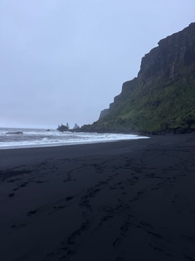 The image depicts the black sand beach of Reynisfjara, near Vik, Iceland. The scene is dominated by a dark, imposing cliff face covered in green vegetation on the upper left. The beach, consisting of black volcanic sand, takes up the right side of the image, with visible footprints. The Atlantic Ocean is in the center, with waves crashing on the shore. The sky is overcast and gray, creating a somber atmosphere. In the distance, rock formations known as Reynisdrangar protrude from the sea. The orientation of the image is horizontal, though it appears to be tilted slightly.