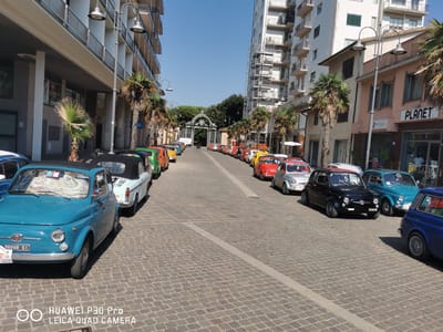 The image shows a street lined with parked Fiat 500 cars in Follonica, Italy. The cars are of various colors including blue, white, red, orange, yellow, and black. They are parked along a cobblestone street on what appears to be a sunny day, with clear blue skies. Buildings with balconies and palm trees line the street. In the background, a large decorative gate is visible. The scene suggests a gathering or exhibition of vintage cars.