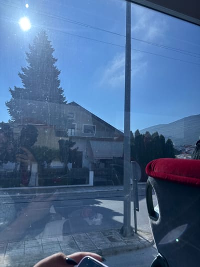 A sunlit, outdoor scene is viewed through a dirty window, likely from a vehicle. A large evergreen tree is silhouetted against a clear blue sky, with the bright sun creating lens flare. In the background, a multi-story house with a dark roof is visible, along with other trees and buildings. A street with a sidewalk and what appears to be parked vehicles is also present. The foreground shows a portion of a car seat with a red headrest, and a person's hand with dark nail polish holding a smartphone. Reflections on the window obscure details and suggest movement, possibly indicating the photo was taken while traveling. The environment appears to be a residential area within or near the city of Kato Nevrokopi, Greece, suggested by the architecture and mountainous backdrop.