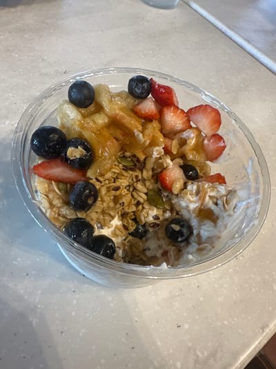 The image displays a clear plastic bowl containing a food item, likely a yogurt or fruit parfait, positioned on a light-colored, speckled countertop. The bowl's contents consist of a white base layer, a substantial quantity of granola comprising oats and various seeds (including flax and pumpkin seeds), and fresh fruit. Specifically, sliced red strawberries and whole dark blue blueberries are distributed across the top. A viscous, golden-brown liquid, consistent with syrup or honey, is drizzled over the fruit and granola, as well as over strips of light-brown, translucent, and irregularly shaped material. The countertop is visible in the foreground and extends into the blurred background. No individuals or specific actions are depicted. The exact location within Medford, United States, cannot be determined from the visual information present in the image.
