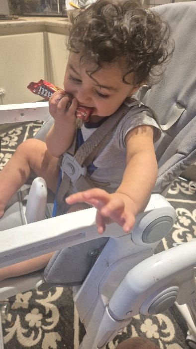 A single infant or toddler, characterized by dark, curly hair, is depicted seated in a light grey and white high chair. The child wears a light-colored t-shirt, possibly grey, with a dark star outline design, and a diaper or brief. A multi-point safety harness, including shoulder straps and a central buckle, secures the child within the chair. The child holds a red-wrapped "m&m's" chocolate bar with both hands, actively biting into the exposed portion of the chocolate. The child's right hand (viewer's left) is slightly extended forward, fingers partially curled, resting on the high chair's white plastic armrest. The immediate environment includes light-colored, possibly beige, kitchen cabinets or wall panels in the background. The floor beneath the high chair features a dark-colored rug with a distinct light-colored, ornate, possibly floral or damask, pattern. The specific location within Melle, Belgium, is not discernible from the visual evidence.