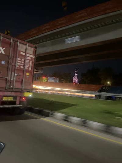 The image depicts a nighttime scene on a highway, likely in or near Sawotratap, Indonesia, given the provided context. In the foreground, the rear of a large red shipping container truck is visible on the left, partially obscuring the road. The truck has its brake lights illuminated. To the right of the truck, a highway stretches into the distance with blurred streaks of light indicating moving vehicles, particularly noticeable are the white light trails of headlights and taillights. The highway is bordered by a grassy embankment on the right, with a concrete barrier in between. An elevated highway structure is prominent in the upper portion of the image, with its underside visible. In the background, silhouetted trees and distant lights suggest an urban or suburban environment. A blurred structure, possibly a statue or monument, is faintly visible in the middle ground to the right, illuminated by red lights at its base. Another vehicle, a dark-colored truck or van, is moving on the highway to the right. The overall impression is of a busy highway environment at night, with the motion of vehicles contributing to the blur and streaks of light.