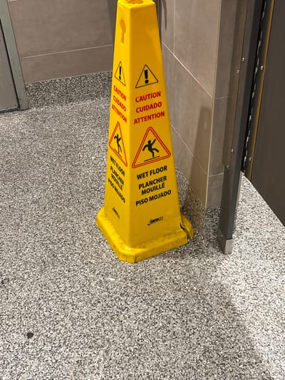 A bright yellow "wet floor" sign stands on a speckled gray floor. The sign has black text and symbols, including warnings in English, Spanish, and French, and a pictograph of a person falling. The sign is positioned in front of a tiled wall and next to a metal post, suggesting it is in a public area such as a restroom or hallway. The floor appears to be a hard, durable material.