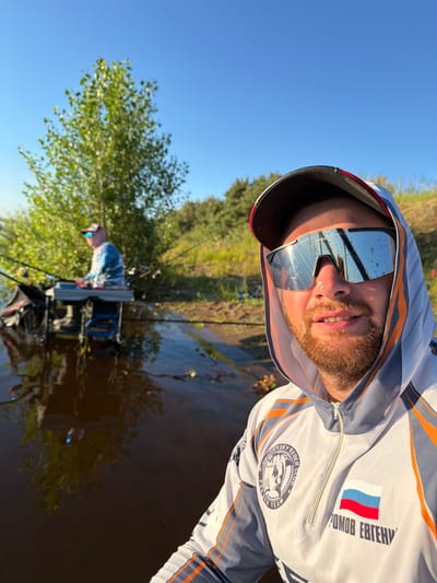A close-up selfie shows a man with a beard and sunglasses, wearing a hooded fishing shirt. His reflection is visible in his sunglasses. In the background, another person is fishing from a chair by the water, with fishing rods set up. The scene appears to be a body of water with trees and vegetation on the bank under a clear blue sky. The man's fishing shirt has an emblem and what appears to be a Russian flag with text. The location is identified as Tarasovo, Russia.