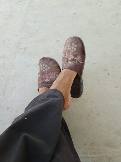A person's legs, clad in dark trousers and brown embroidered slippers, are resting on a light-colored, textured surface. The slippers appear to be made of a fabric material, with a floral embroidery pattern featuring pink and light brown flowers. The surface resembles concrete or a similar paved outdoor area, with scattered small debris. The lighting suggests daytime, possibly outdoors, given the even illumination and the dry, somewhat dusty appearance of the ground and slippers. The scene is a simple, casual pose, with the feet crossed. There is no visible text. The location is identified as Muborak, Uzbekistan.
