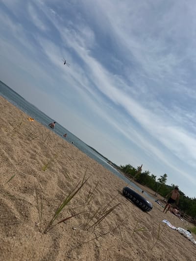 The image depicts a beach scene with sand in the foreground, leading to a body of water. The sky is blue with wispy white clouds. A dragonfly is visible in the sky. Several people are in the water, and some are on the beach. One person is running on the sand, and a black inflatable tube is lying on the sand. Trees and foliage are visible in the background. The scene suggests a warm, sunny day at a lake or sea, possibly in Aiyrtau, Kazakhstan, given the sandy terrain and vegetation typical of the region.