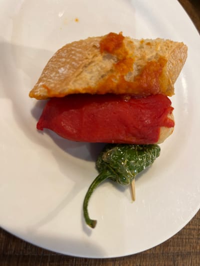 A white plate with a red pepper, a green pepper, and a piece of bread with red sauce on top. The red pepper is on top of the bread and the green pepper is underneath. The green pepper is on a toothpick. The scene appears to be on a table. The coordinates 43.324, -1.985 correspond to the location of Donostia, Spain.  This is a typical example of the common Spanish dish "pintxos" (also called "pinchos") served in Donostia.