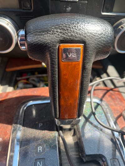 The image shows a close-up of a car's gear shifter and dashboard, which appears to be inside a vehicle located in Aqköl, Kazakhstan. The gear shifter is made of black leather with a wood-grain accent and a "V8" emblem. The dashboard features two circular gauges and a panel with gear indicators "P," "R," and "N." Wires are visible in the background, suggesting the car's interior is somewhat exposed or undergoing maintenance. The lighting indicates it is daytime, with sunlight casting reflections and dust particles on the surfaces.