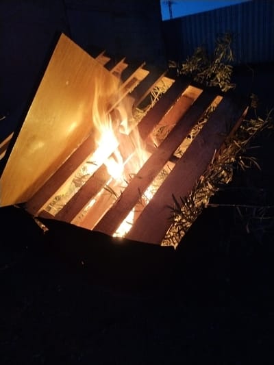 The image captures an outdoor scene at night, focusing on a contained fire. Bright orange and yellow flames emanate from the center, illuminating the immediate surroundings. The fire is burning within a square-shaped structure constructed from dark wooden slats, resembling a makeshift fire pit, possibly fashioned from a discarded pallet or crate. A larger, lighter-toned wooden board leans into the structure from the upper left, reflecting the fire's glow.
The fuel for the fire appears to include various pieces of wood and bundles of dried foliage or branches, some of which are visible along the right edge of the wooden enclosure and intermingled with the burning material. Given the context of Lo Ferro, Spain, these branches likely belong to olive trees, common in the region.
The time of day is clearly night, as indicated by the dark, indistinct ground and background, with the fire serving as the primary light source. In the background, dark shapes suggest a building or fence, and a faint blue light can be seen towards the top right, possibly a distant streetlamp or part of the night sky. No people are visible in the image, nor is there any discernible text. The scene depicts a practical outdoor burning activity, potentially for warmth, light, or disposal of organic waste.