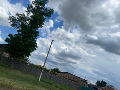 The image depicts a cloudy sky with some patches of blue, above a scene that appears to be in the Tagarogsky district of Russia. In the foreground, a sloped grassy area leads up to a wooden fence topped with a yellow railing. Behind the fence, there are buildings with dark roofs and some trees. A utility pole with wires runs diagonally across the frame. The overall impression is one of a somewhat overcast day in a semi-rural or industrial area.
