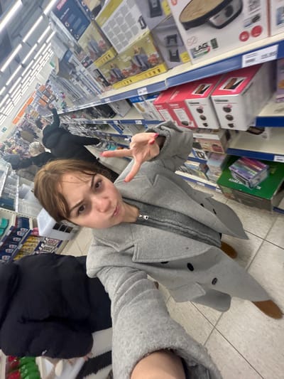 A selfie depicts a woman in a gray coat, making a peace sign while standing in a store aisle. Shelves stocked with various products line the left side of the frame. Other people are visible further down the aisle. The woman is wearing brown shoes and a gray sweater under her coat, which has black buttons. The fluorescent lighting of the store illuminates the scene.