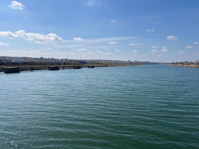 The scene depicts a wide view of the Poarta Albă–Midia–Năvodari Canal in Medgidia, Romania. The foreground consists of dark green-blue water with a rippled surface. A multi-pier concrete structure, consistent with a low-profile bridge or conduit, extends from the left bank, partially over the canal. Both the left and right banks are lined with dry, brown vegetation. The city of Medgidia occupies the distant left and center horizons, characterized by numerous residential buildings with varied roof colors and some industrial structures on elevated terrain. A prominent arch bridge spans the canal in the far distance, slightly right of center. The sky is clear blue with scattered white cumulus clouds; a faint contrail is visible in the upper right quadrant. The environment suggests bright daylight conditions. No human or animal subjects are visible within the frame.