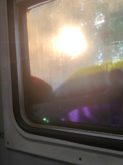 The image shows a view through a dirty train window. The sun is shining brightly through the window, creating a glare and making it difficult to see details inside the train. There are some blurry shapes visible through the window, which could be people or objects. The exterior of the window frame is visible, and it appears to be made of white plastic. The interior of the train is dark, with some purple fabric visible at the bottom of the frame. A small green light is also visible on the window. There are no explicit clues to indicate the location in Hlevakha, Ukraine.