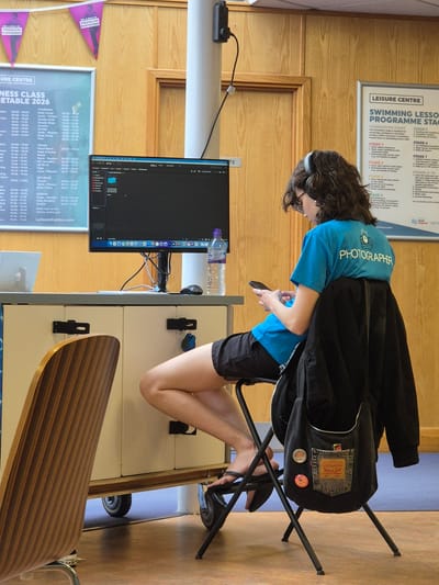 A woman with curly dark hair sits on a black folding chair, facing left, and looking down at a cell phone in her hands. She wears a blue t-shirt with the word "PHOTOGRAPHER" printed on it, along with black shorts and black shoes. She has headphones around her neck and a black jacket hanging on the back of the chair. The chair is positioned next to a white cabinet on wheels. A computer monitor is on the cabinet, displaying a dark screen. A water bottle is also on the cabinet. In front of the cabinet is a desk with a striped wooden top.
Behind the woman and the cabinet, there is wood paneling on the wall. Hanging on the wall are two posters. One has "LEISURE CENTRE" in large letters and the second has "URE CENTRE" and "NESS CLASS" printed on it. Additionally, above the wood paneling, there is another poster with the words "LEISURE CENTRE", "SWIMMING LESSO", and "PROGRAMME STAC" printed on it. The location is the Breckland District in the United Kingdom.