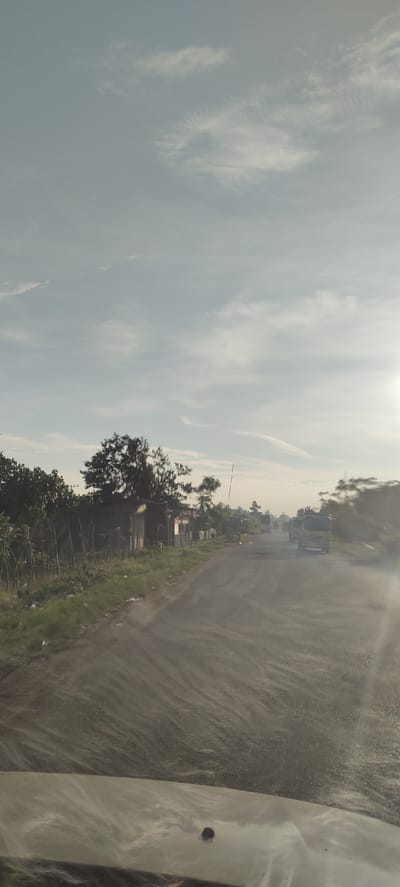 A wide-angle, eye-level shot captures a dusty, rural road under a hazy, pale blue sky with wispy white clouds. The road, which appears to be unpaved or undergoing construction, stretches into the distance, with tire tracks creating a striped pattern on the surface. To the left of the road, a line of lush green vegetation borders a small, dilapidated shack. Beyond the shack, several trees with dense canopies rise, casting shadows. In the middle ground, a few trucks are visible on the road, their forms softened by the atmospheric haze. The sun, positioned on the right side of the frame, casts a bright glare that washes out details in the distance. The foreground shows the hood of a vehicle, suggesting the image was taken from inside a car. This scene is likely located in the city of Meunasahmesjid, Indonesia, given the context.