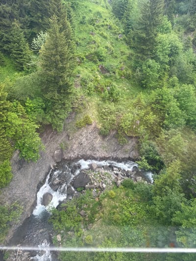 An aerial, high-angle, medium shot captures a winding, narrow river flowing through a rocky gorge. The river is a mix of white rapids and darker, deeper pools, with numerous large rocks scattered throughout its course. The gorge walls are steep and rocky, with a variety of green foliage growing on them, including trees and bushes. The surrounding landscape is a lush, green mountainside covered in trees and grass. The scene appears to be located in a natural, mountainous environment, consistent with the terrain of Hasliberg Reuti, Switzerland. The lighting suggests it is daytime, possibly overcast, with diffused natural light illuminating the scene. There are no people or man-made structures immediately visible.