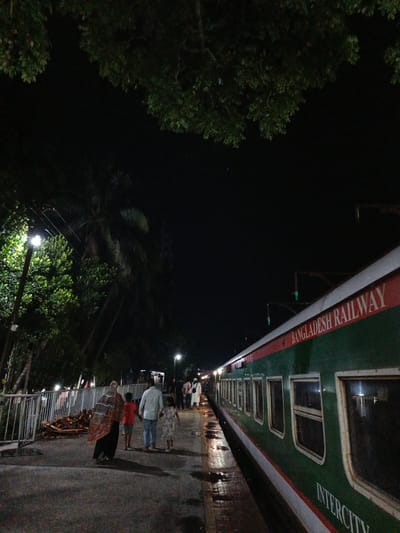 The image depicts a night scene at the Rajbari Railway Station in Rajbari, Bangladesh. A green and red Bangladesh Railway train is parked at the platform. Several people are visible on the platform. In the foreground, a family consisting of two adults and two children are walking away from the camera. The adult on the left, possibly a woman, is wearing a colorful patterned headscarf and a dark outfit. Beside her, a child in an orange shirt walks, followed by an adult male in a white shirt and blue jeans, holding the hand of a child in a light-colored dress. To the right of the train, the platform is wet, reflecting the lights. In the background, more people are seen walking on the platform, and some lights illuminate the area. Trees and foliage are visible on the left side of the image, with a street light illuminating the trees and a section of the platform. The sky is dark, indicating it is nighttime.