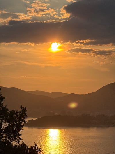 The photograph captures a sunset over a coastal landscape, likely in or near Čelobrdo, Montenegro. The sun, partially obscured by clouds, casts a warm, orange glow across the sky and water. The sun's rays create a shimmering reflection on the surface of the sea, which fills the lower portion of the frame. In the middle ground, silhouetted hills and mountains recede into the distance. A town or settlement is visible nestled along the coastline, with buildings faintly discernible against the dark hills. In the foreground, the dark silhouettes of trees and foliage frame the left side of the image. The scene is devoid of any human presence or discernible activity. The overall impression is one of natural beauty and tranquility.