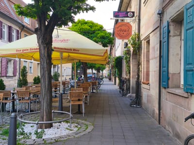 This image depicts an outdoor seating area of a cafe or restaurant, likely located in Erlangen, Germany, as suggested by the presence of the "Lederer Biere" sign, a well-known brewery from Erlangen. The scene is set along a cobblestone sidewalk lined with buildings. In the foreground, a large yellow umbrella provides shade over several tables and chairs. A mature tree with a thick trunk stands prominently in front of the umbrella, its branches extending upwards. The tables are set with chairs, some with woven seats and backs, and metal legs.
Further down the sidewalk, more tables and chairs are visible, suggesting a busy establishment. Several bicycles are parked along the side of the building, indicating their use by patrons or locals. The building facades appear to be made of stone, with windows featuring blue shutters. Two signs are visible hanging from the building: one for "Lederer Biere" with a logo, and another octagonal sign for "Anna's Cafe - Bar - Bistro" with "EST 2022" printed on it. The environment appears to be a public space, with potted plants and greenery adding to the ambiance. The overall impression is of a charming, European street scene during the daytime.