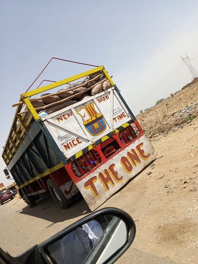 A large goods transport truck is depicted on a road in Toro, Nigeria. The truck, viewed from its rear-right side, is heavily loaded with numerous large, irregularly shaped, light-colored sacks, likely containing agricultural produce, stacked above its bed and secured by wooden poles and red ropes. The truck's rear features a white upper section, a red lower section, and a yellow frame around the cargo area. Decorative text includes "NEW MOD NICE," "TIME," and a centrally painted FC Barcelona crest. The phrase "THE ONE" is prominently displayed in yellow-orange letters on a white bumper flap. Smaller text mentions "DAWUSHA ENTERPRISE" with a phone number and "BEST BY M. MUSTAFA NPA. JUT" (partially obscured). Blue decorative lights are visible through a red grate at the lower rear. The vehicle is on a light-colored paved road. To its immediate right is a dry, sandy shoulder with sparse vegetation. In the distant right background, a tall metal electricity pylon stands against a clear, bright blue sky. Another vehicle is partially visible on the road behind the truck. The image is captured from a tilted perspective, originating from inside another vehicle, with a side-view mirror occupying the lower foreground, reflecting a person wearing a white garment.