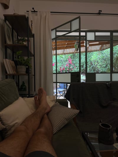 The image shows the legs of a person lying on a couch. The person is wearing shorts and has their feet crossed. They are relaxing on a green couch in a living room. In the background, a large, multi-paned window looks out onto a patio with a wooden pergola. Through the window, one can see a lush, green garden with pink flowers and a patio set. On the right side of the image, a dark gray blanket is draped over a couch, obscuring the lower half of the figure sitting on it. A small table with a ceramic mug on top is positioned in front of the blanket-covered couch. 
The image is likely taken from a home in the city of Alon HaGalil, Israel, as indicated by the provided coordinates (latitude: 32.755, longitude: 35.219).