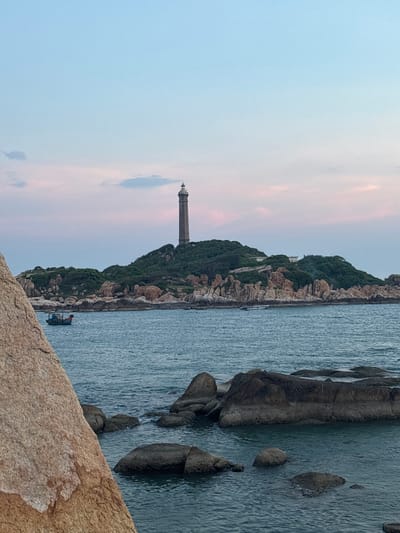 The image depicts a lighthouse on a rocky island off the coast of Thuận Thành, Vietnam. The lighthouse is a tall, cylindrical structure with a cap and a lantern room at the top. It is situated on a hill covered in dense green vegetation and surrounded by numerous large, light-brown boulders. The sea in the foreground is a deep blue, with several dark, rounded rocks emerging from the water. A small, dark blue and red fishing boat is visible on the water to the left of the island. The sky above is a gradient of soft blues and pinks, suggesting either sunrise or sunset. The foreground is dominated by a large, rough-textured, light-brown boulder that partially obscures the view of the sea and the island. The scene is serene and natural, emphasizing the stark beauty of the coastline.