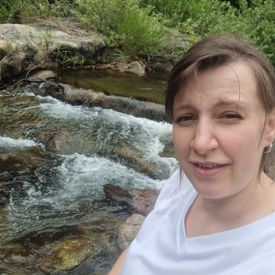A medium shot selfie shows a woman with brown hair and a light complexion, wearing a white t-shirt, standing in front of a flowing stream. The stream is rocky, with clear water moving over submerged stones. Lush green foliage and larger rocks line the bank behind the stream. The woman's face is angled towards the camera, with a slight smile and her eyes looking at the viewer. She has a small silver hoop earring in her visible ear. The background suggests a natural, outdoor setting, possibly a park or a hiking trail. The lighting appears to be natural daylight.
