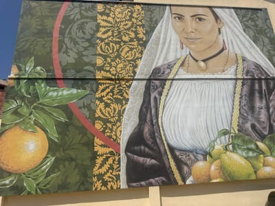 The image is a mural painted on the side of a building. It depicts a woman wearing traditional attire, including a white veil, a dark patterned dress, and elaborate jewelry. In the foreground, there are clusters of ripe oranges and lemons on branches. The mural incorporates patterned backgrounds in green and gold. To the upper left of the mural, there is a signature that appears to read "Tatte '21". The mural is located in Muravera, Italy.