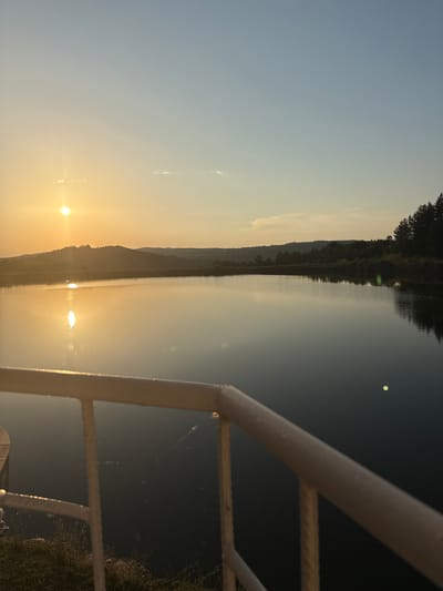 A wide, still body of water reflects the warm glow of a setting sun. The sky above transitions from a pale yellow near the horizon to a soft blue higher up. In the distance, rolling hills are silhouetted against the light, with a line of dark trees along their crest. In the foreground, a white railing, likely part of a balcony or deck, partially obstructs the view of the water. The sun, a bright orb, casts a shimmering reflection on the water's surface. The overall scene evokes a sense of tranquility and natural beauty, possibly at dusk. The location is identified as Skalsko, Bulgaria.