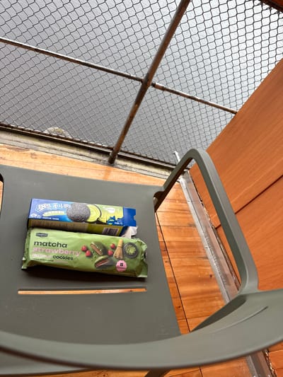 The image captures an outdoor scene, likely a balcony or deck in Lappeenranta, Finland, with a focus on two packages of cookies. A dark grey, modern-style chair occupies the lower left portion of the frame, angled towards the right. Resting on the chair's seat are two cookie packages:
1. **Top Package:** A blue cardboard box, appearing to be an Oreo brand product with "Matcha" flavor, featuring Chinese characters and an illustration of a black cookie with green filling.
2. **Bottom Package:** A green flexible plastic pouch from the "GoodBurry" brand, clearly labeled "matcha strawberry flavour cookies," displaying images of green-coated cookies with red strawberry centers. It indicates "8 cookies" and "128g."
The chair rests on a light brown wooden plank floor, which shows some wear and dust in the gaps. Beyond the wooden floor, a sturdy metal railing runs horizontally, followed by a grey chain-link fence that spans the upper two-thirds of the image. Through the fence, a large, calm body of grey water is visible, consistent with the appearance of Lake Saimaa under an overcast sky. The sky itself is a uniform grey. On the far right, a vertical section of a reddish-brown wooden wall or paneling is present, suggesting an adjacent building or privacy screen. No individuals are present in the scene.