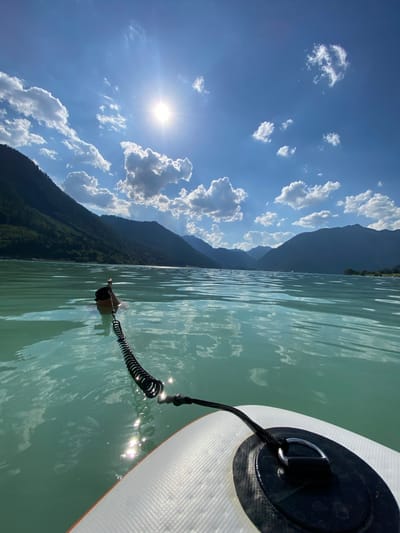 The image depicts a person on a paddleboard in a lake, with mountains in the background. The sun is shining brightly, and there are some white clouds in a clear blue sky. The water is calm and reflects the sunlight. The person is wearing a dark top and appears to be paddling, though their face is not visible. The paddleboard is white with an orange trim and has a black leash attached to a D-ring. The scene is set in a mountainous region, with lush green trees covering the slopes of the mountains. The time of day is likely midday, given the bright sunlight. The weather is sunny and pleasant, ideal for outdoor activities. The overall mood of the image is serene and relaxing, capturing the enjoyment of a day on the water. The location cues, such as the type of mountains and the clear lake, suggest a natural setting in a European alpine region. No visible text is present in the image.