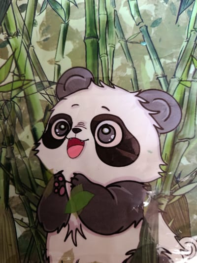 This is a close-up, slightly angled photograph of a cartoon panda against a background of stylized green bamboo stalks. The panda is depicted in a cheerful, playful manner. It has large, expressive eyes with prominent black eye patches, a small nose, and a wide, open mouth showing a pink tongue and red gums, giving it a happy and excited expression. The panda has black ears and black markings on its arms and legs. It is holding a small green leaf in its paws, suggesting it might be eating or playing with it. The bamboo stalks are depicted in various shades of green with dark outlines, creating a sense of depth and a forest-like setting. The overall image has a bright, cartoonish aesthetic, and appears to be a sticker or part of a larger printed design, possibly on a plastic surface, as indicated by some visible reflections and creases in the material. The time of day and weather are not discernible. The location context provided is Smarhon, Belarus, but there are no specific visual cues in the image to confirm this location. There is no visible text in the image.