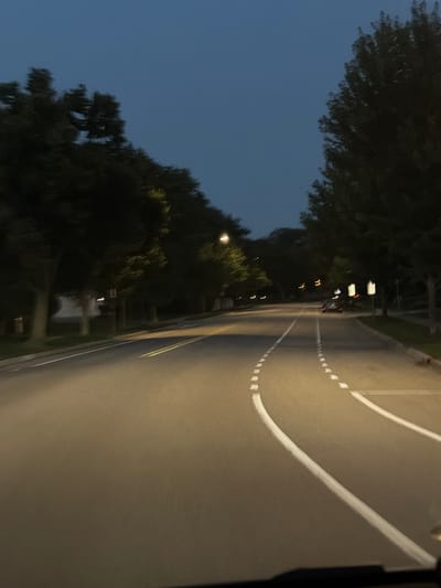 The image shows a street scene in Vernon Hills, United States of America, during dusk or early evening. A paved road with yellow and white lane markings is visible, with a car in the distance driving away from the camera. The road is lined with trees on both sides, casting shadows and creating a somewhat dim atmosphere. Streetlights are present, illuminating parts of the road. The sky is a deep blue, indicating the transition from day to night. The perspective suggests the photo was taken from inside a vehicle.