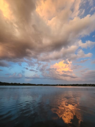 This is a photograph of a body of water, likely a lake, on a partly cloudy day. The sky is filled with dramatic clouds, illuminated by the setting sun with hues of orange, pink, and yellow. The water reflects the sky and the distant tree-lined shore, creating a mirror-like effect. The far bank of the water is lined with a dense strip of trees, behind which are visible houses and structures, suggesting a residential area. The water is calm with gentle ripples, and there are no people or boats visible in the scene. The overall impression is one of serene natural beauty and the tranquility of a late afternoon or early evening. This scene could be found at a location such as Lake Virginia or another of the many lakes within Winter Park, Florida, known for its picturesque lakes and abundant natural beauty.
