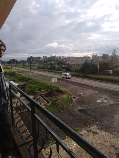 The image presents an elevated, wide-angle view of a semi-urban environment within Njoro ward, Kenya, under an overcast sky.
In the immediate foreground, the left side is occupied by the partial figure of an individual, from the chest up, wearing a dark beanie with a colorful pattern and a dark garment, possibly a jacket or shirt. A strap is visible over their left shoulder. The person's face is turned towards the camera. A dark, ornate metal balcony railing extends across the lower portion of the frame. Below the railing, hexagonal paving stones are visible, some appearing damp or stained, along with a translucent, coiled object on the right side.
The midground features an unpaved, two-lane road with visible ruts and standing water, indicating recent moisture. A white passenger van, typical of a matatu, travels on the road from right to left. Wooden utility poles with multiple power lines run parallel to the road. To the left of the road, a small, light-colored sign with the text "SUKAKA SUPERMARKET" is posted adjacent to a pile of disturbed earth and debris. Green grass and scattered vegetation flank the road. Another distant vehicle is faintly visible further along the road.
The background reveals a mix of sparse trees and various buildings, including multi-story structures under construction or incomplete, characterized by visible concrete blocks. Other structures appear to be painted residences or commercial establishments. The horizon is defined by a dense line of trees. The sky is predominantly covered by grey, stratiform clouds, suggesting an overcast condition.