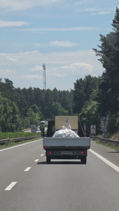 The media file shows a truck driving on a highway in Calau, Germany. The truck is loaded with a large wooden crate and a white tarp-covered object. The scene is set during the daytime, under a clear blue sky with some scattered clouds. Lush green trees line the sides of the highway, suggesting a rural or suburban environment. Several road signs are visible, including parking signs, a speed limit sign of 60 km/h, and a triangular warning sign indicating a hazard ahead with "2 km" written below it. A construction crane can be seen in the distance on the left side of the image. The truck has a license plate with the number "1822498". The overall impression is of a bright, sunny day with a clear view of the road and surrounding landscape.
