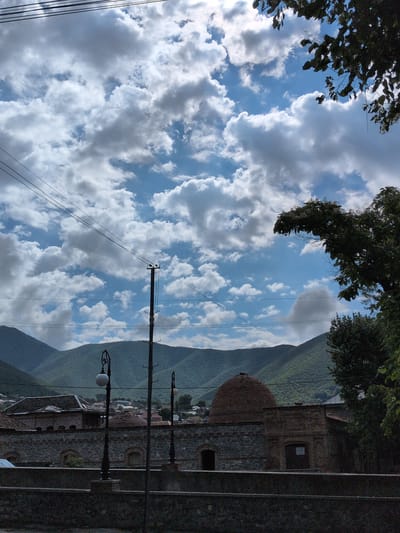 This media file captures an outdoor scene in Şəki, Azerbaijan, likely during the daytime under cloudy skies. The image is oriented vertically, with the top of the frame showing dark green foliage from trees against a bright blue sky dotted with fluffy white clouds.

Below the foliage, a mountain range covered in lush green vegetation dominates the background. The slopes of the mountains are densely forested, suggesting a natural or semi-natural landscape. In the mid-ground, architectural elements of a building are visible, constructed with rough-hewn stone and brick. A prominent feature is a rounded, reddish-brown dome, possibly part of a historical structure like a mosque or a mausoleum. The building has dark-framed windows and arches.

In the foreground and mid-ground, there are also lampposts with ornate fixtures, typical of older street lighting. Electrical wires and a utility pole are visible, crossing the sky and indicating the presence of infrastructure. The overall impression is one of a picturesque setting where nature meets human architecture. The weather appears to be fair, with the clouds suggesting a possibility of rain or a dynamic sky. There is no visible text in the image.