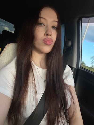 The media file is a selfie of a young woman taken inside a car, likely in Gavrilovka, Russia. She has long, dark brown hair that falls over her shoulders and chest. She is wearing a white t-shirt and a silver necklace. Her makeup is visible, with emphasis on her eyes and lips, which are slightly parted. The image is taken at an angle, showing a portion of the car's interior, including a seatbelt and the roof lining. Outside the car, the sky is visible through the sunroof and window, appearing to be daytime with some trees and foliage in the background. The overall mood appears casual and relaxed, possibly during a car journey. There is no visible text in the media file.