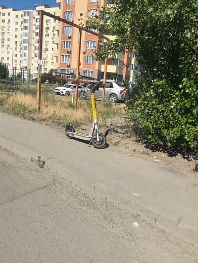 A yellow and silver electric scooter stands on a dirt path next to a curb. The scooter has "WHOOSH" written vertically in yellow on its stem, with "PE4190" below it. Behind the scooter, a rusty metal frame structure is present. In the background, there are apartment buildings with many windows and air conditioning units. A white car and a silver car are parked on the street. Green trees and dry grass are visible, suggesting an outdoor setting. The sun appears to be shining, indicating a bright, daytime scene. The area seems to be a residential neighborhood in Verkhnetemernitsky, Russia. There is no visible activity of people or events taking place. The overall mood is neutral, depicting a typical urban environment.