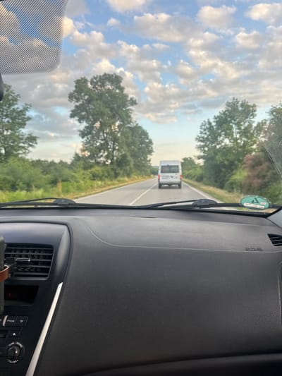 The image was taken from inside a vehicle driving on a rural road, likely near Draganovtsi, Bulgaria. The sky is partly cloudy with a mix of blue sky and white clouds. Lush green trees and vegetation line both sides of the asphalt road. In the distance, a white van is driving ahead on the same road. The foreground shows the interior of the vehicle, including the dashboard and windshield wipers. A circular sticker with green and white markings is visible on the lower right side of the windshield.